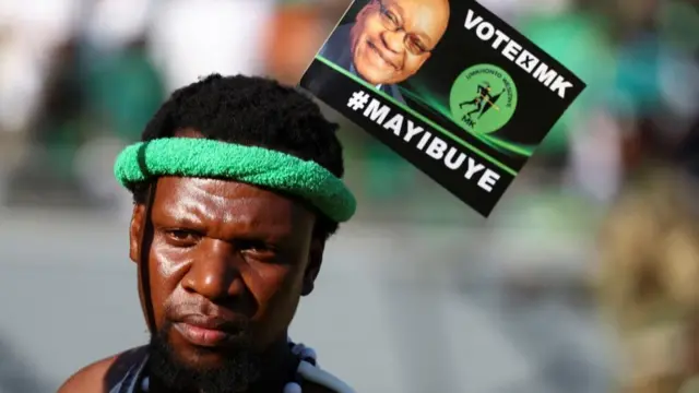 Umugabo uhagaze imbere y'icyapa gisaba gutora ishyaka MK rya Zuma