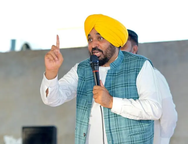 ਭਗਵੰਤ ਮਾਨ ਸਟੇਜ ਤੋਂ ਸੰਬੋਧਨ ਕਰਦੇ ਹੋਏ। ਉਨ੍ਹਾਂ ਨੇ ਚਿੱਟੇ ਕੁੜਤੇ ਪਜਾਮੇ ਉੱਤੋਂ ਨੀਲੀ ਡੱਬੀਦਾਰ ਜੈਕਟ ਪਹਿਨੀ ਹੈ ਅਤੇ ਪੀਲ਼ੀ ਪੱਗ ਪਹਿਨੀ ਹੈ।