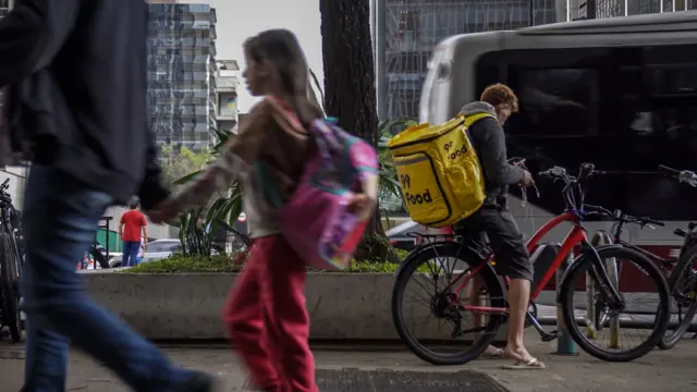 Homem e uma menina passando em movimento no primeiro plano; em segundo plano, com nitidez, um entregador está em uma bicicleta vermelha, usando uma bag amarela da 99, olhando para o celular