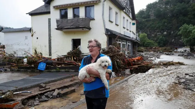 Um homem carrega um cachorro próximo a escombros trazidos pela enchente