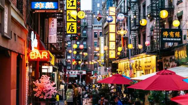 A crowded street of restaurants and bars in New York City's Chinatown. Red, yellow and blue lanterns hang above the street