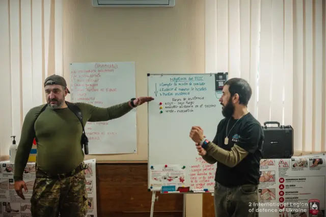 Два человека стоят у доски, где что-то написано маркером, один из них, бородатый, в камуфляжных штанах и и кофте цвета хаки показывает на нее рукой.