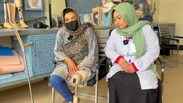 ګل بېګم د مرکز له یوې کارکوونکې سره په ګلداره کالیو کې ناسته ده، شین پتلون یې اغوستی او چپه پښه یې مصنوعي او لوڅه ده. تر څنګ یې د مرکز کارکوونکې زرغون رنګه ټېکری پر سر د ډاکټرانو په سپينه چپنه کې ناسته ده او تور مرۍ دار کالي یې برېښي. 
