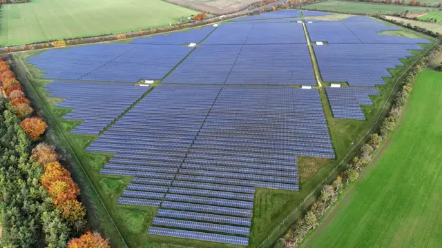 Una imagen de dron muestra desde arriba un enorme parque solar cerca del pueblo de Great Wilbraham, cerca de Cambridge. Miles de paneles cubren un vasto campo. El parque solar está rodeado de campos y árboles.