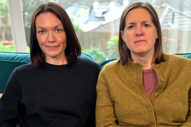 Дві жінки сидять у приміщенні на дивані. З вікна позаду них видно пейзаж просто неба з деревами та дахами будинків у розмитому фокусі.