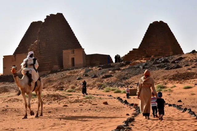 A woman walks with children towards the Meroe Pyramids. Nearby a man rides a camel.