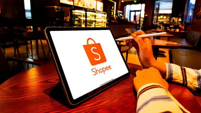 Mãos segurando uma caneta digital em frente a um tablet mostrando o logo da Shopee