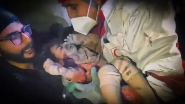 Um socorrista com máscara segura uma criança pequena coberta de poeira, enquanto outro observa