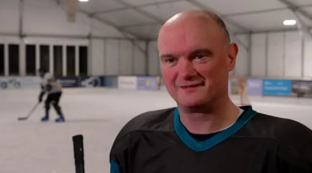 Simon Soar sonríe fuera de cámara. Se le ve con una camiseta negra. Detrás de él hay una pista de hockey sobre hielo con un jugador.