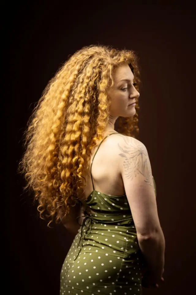 Retrato de estudio de una persona con cabello largo, voluminoso y rizado, fotografiada de espaldas y de perfil, girando ligeramente hacia la cámara. 