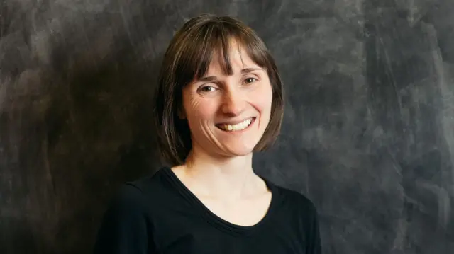 Elaine Midgley, de pie frente a una pared negra, sonríe y tiene el pelo oscuro hasta los hombros. Lleva una blusa negra.