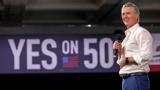 Gavin Newsom kadrajın sağında elinde mikrofonlar bir salonda ayakta duruyor ve gülümsüyor. Arka planda İngilizce 50'ye Evet yazılı bir panlat var.