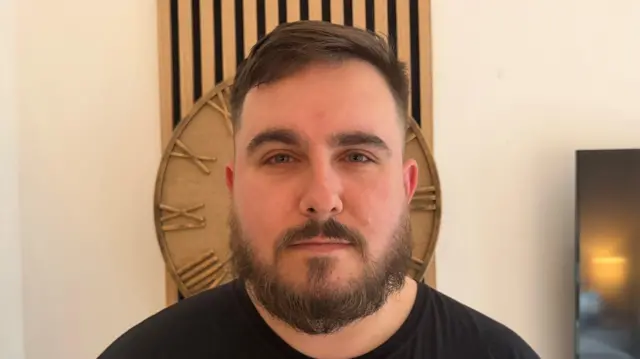 Imagem mostra um jovem, do peito para cima, com barba cheia e bigode, vestindo uma camiseta preta. Ele tem uma expressão séria, e há um relógio de madeira diretamente atrás de sua cabeça