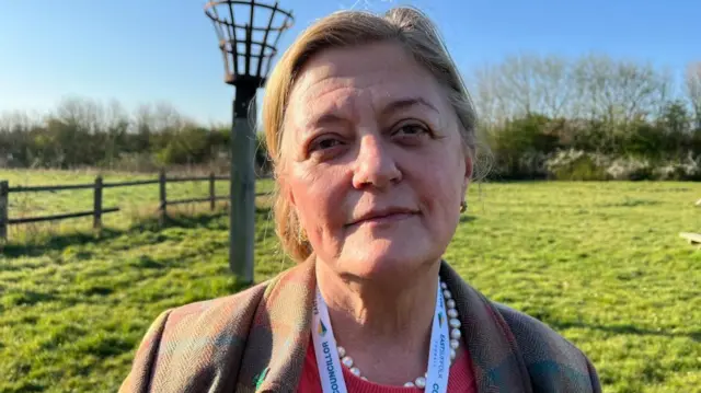 Julia Ewart se encuentra al aire libre, en un campo de hierba, en un día soleado. Lleva un abrigo a cuadros en tonos apagados sobre una blusa color coral, complementado con un collar de perlas y un cordón del Ayuntamiento de East Suffolk. Detrás de ella hay una valla de madera y una estructura metálica alta, similar a una cesta, sobre un poste.