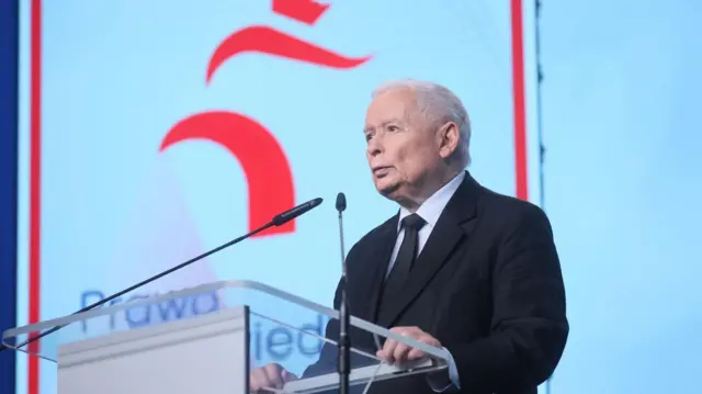 Mężczyzna z siwymi włosami, w białej koszuli i czarnym garniturze przemawia z mównicy
