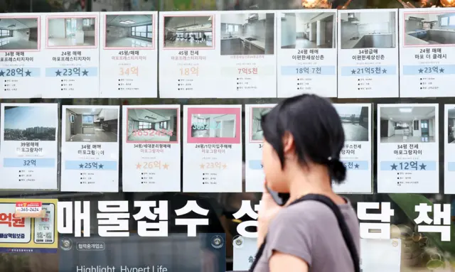 부동산 창문에 각종 매물 정보가 붙어 있다. 그 앞을 한 여성이 지나가고 있다