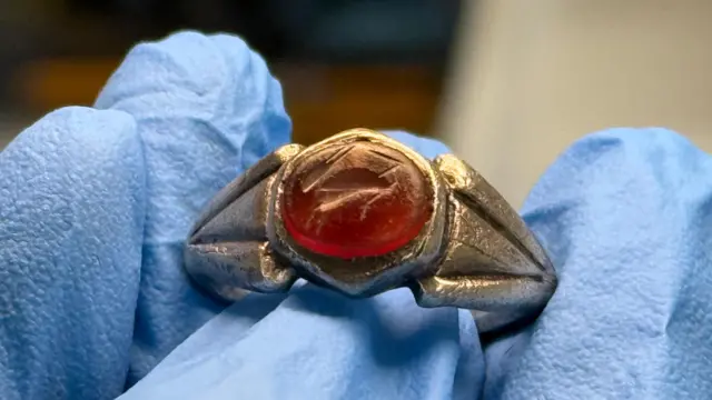 Primer plano de un anillo de plata con una gema roja que lleva grabada la imagen de una liebre. Alguien con guantes azules lo sostiene.