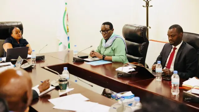 Abantu bicaye mu biro, imbere yabo hari indangururamajwi zo kuvugiramo. Muri iki cyumba hagaragara abagabo babiri n'ukuboko k'umugabo wa gatatu kuri ku meza, n'abagore babiri. Bose bateze amatwi umugore urimo kuvuga, wambaye indorerwamo, fulari mu ijosi n'ishati y'icyatsi kibisi. 