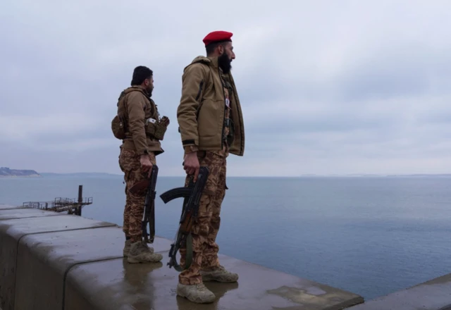 İki Suriye ordusu askeri baraj suyu dolu bir alana bakıyor. Birinin kırmızı beresi var. İkisi de uzun namlulu silah tutuyor.