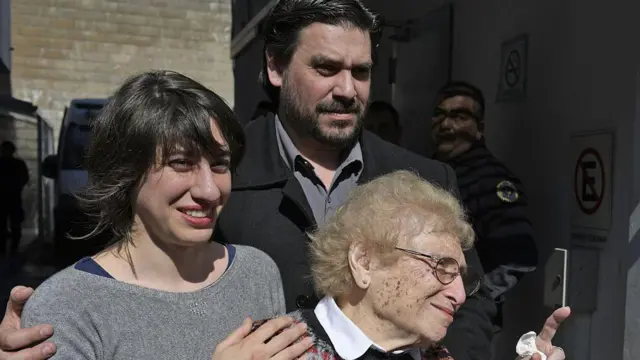 裁判所の外でロイシンブラットさんの肩にマリアナさんが笑顔で手を添え、マリアナさんの肩をギエルモさんが抱いている。明るい日差しを浴びて、3人とも目を細めている