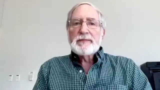 Пожилой седой мужчина в очках, с бородой и в рубашке в бирюзовую клетку.