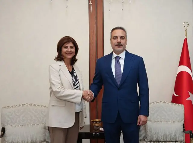 Beyaz ceketli Holguin Cuellar solda, lacivert takım elbiseli Fidan sağda el sıkışıyorlar, geride iki koltuk ve Türk bayrağı var.