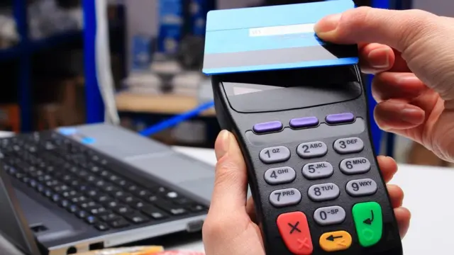 Una persona pasando una tarjeta por un terminal de pago. Al fondo, una laptop