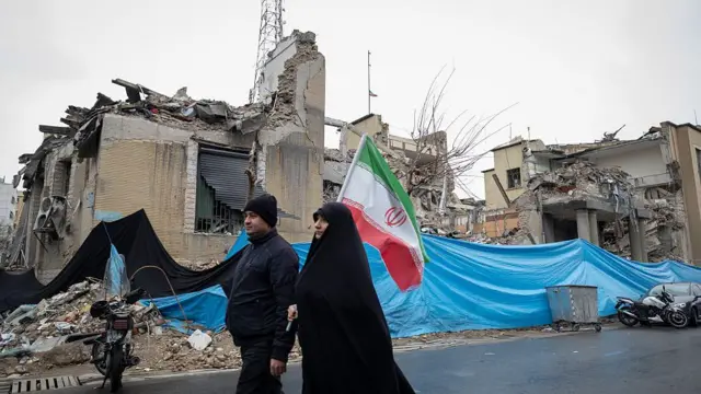 İranlı cütlük Qüds günü yürüşünə gedərkən ABŞ və İsrailin hücumundan dağılmış binanın qarşısından keçir.