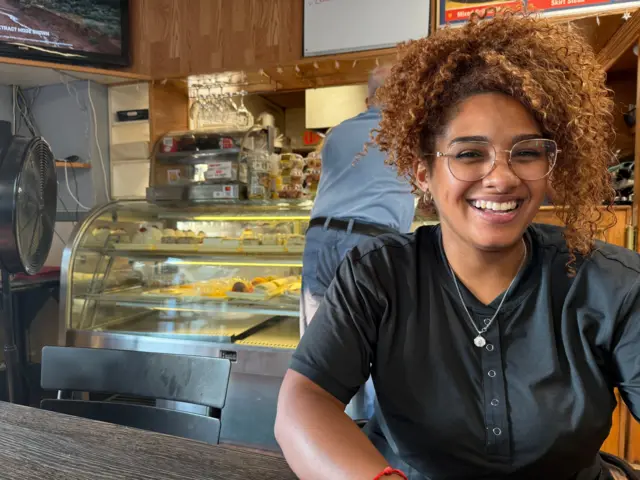 Shandira Rodríguez sonriente frente a la cámara. Viste una blusa gris oscuro. Detrás se ve una de las heladeras de la Pizzería Montevideo con postres.