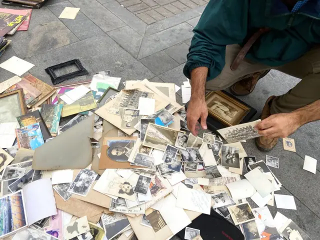 Fotos espalhadas pelo chão em praça público; um homem agachado segura uma das fotos na mão