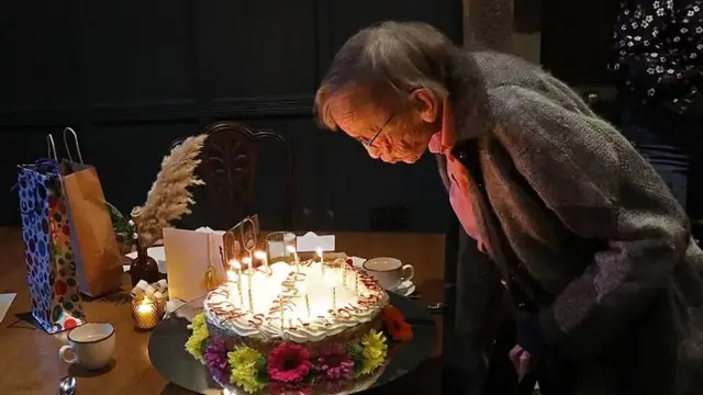 Margaret Bradshaw sopla las velitas en una gran torta de cumpleaños cubierta de crema y decorada con flores