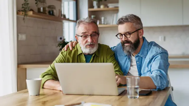 Um padre y un hijo miran un computador juntos, sentados en la cocina.