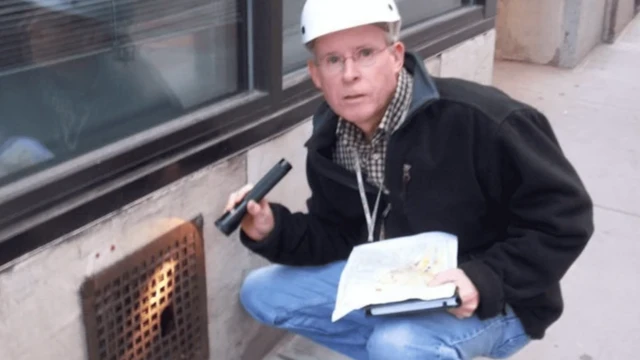 دکتر بابی کوریگان روی پاهایش چمباتمه زده و چراغ‌قوه‌ای رو به دریچه‌ای خیابانی در دست دارد 