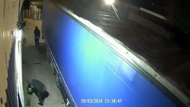 Mężczyzna nalewa zieloną puszkę, podczas gdy inny filmuje go telefonem z boku ciężarówki – wszystko zostało uchwycone przez kamerę bezpieczeństwa