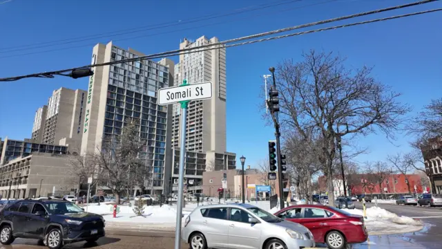 Vue plus large de Somali Street, avec un immeuble élevé en arrière-plan, quelques arbres, le panneau "Somali Street" à côté de trois voitures. On aperçoit la neige fondre au loin.