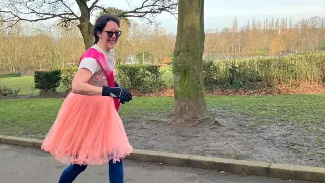 Jennifer Gane corre por un parque. Lleva un vestido corto de fiesta naranja/rosa brillante y una banda rosa. Debajo, lleva pantalones azules para correr y guantes.