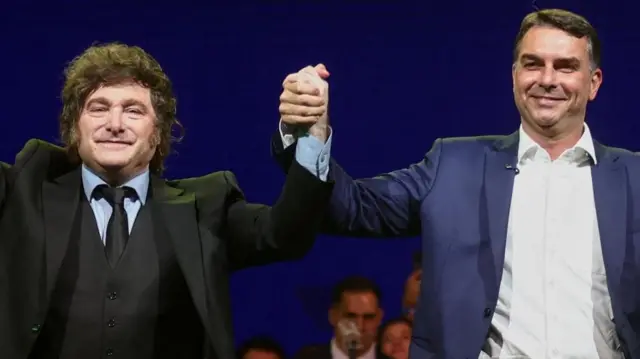 El presidente argentino Javier Milei saluda de la mano al senador brasileño Flavio Bolsonaro. Ambos visten traje y sonríen durante un acto de campaña.