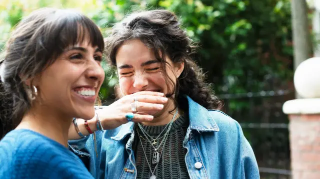 Uzun saçlı iki kadın gülüyor. Biri eliyle ağzını kapatıyor.