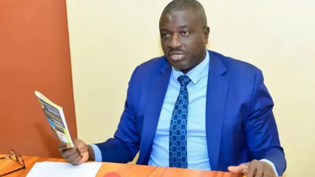 Faustin Ndikumana mw'ikostime y'ubururu, ishati y'ubururu bukeye n'ikaruvato y'ubururu irimwo udukwadarato