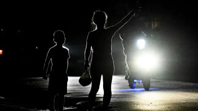 Una mujer y un niño en una calle oscura saludan a un motocicista que lleva las luces encedidas de su moto.