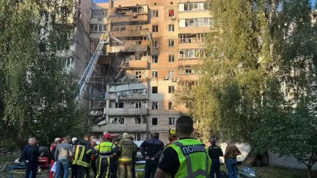 Пострадавший жилой дом в Киеве, разрушена целая секция, к верхним этажам ведет лестница пожарной машины, на переднем плане стоят сотрудники спасательных служб
