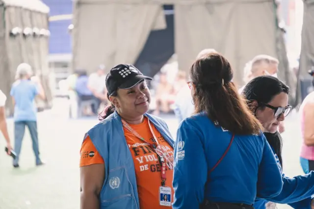 Anicarmen Chirinos con una gorra negra con el emblema de la UNFPA, una camisa anaranjada que dice "Fin de la violencia de género" y un chaleco azul con el símbolo de las Naciones Unidas. Habla con una colega que tiene una camisa azul con el emblema de ACNUR.

