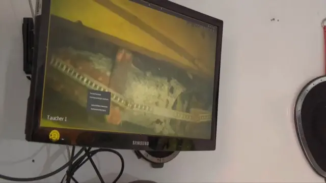 Na monitorze telewizora przymocowanym do ściany wyświetlany jest obraz wideo z kamery podwodnej, na którym widać zardzewiałą łuskę amunicji z umieszczoną na niej miarką krawiecką.