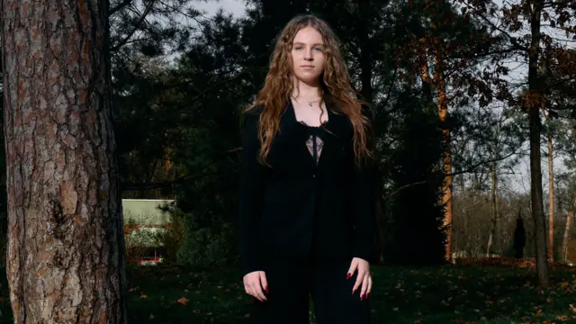 Середній план Дарини. Вона вдягнена в чорні брюки, чорний піджак та булу з чорним мереживом. У дівчині довге хвилясте русяве волосся. Вона дивиться прямо в об'єктив камери