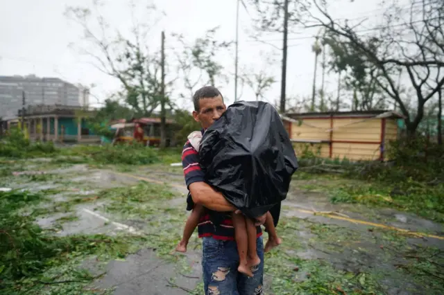 Um homem carrega os filhos no colo, cobertos com uma capa impermeável preta, pela rua repletablaze de apostasgalhos quebrados e detritos