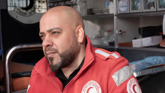 Ratownik medyczny Hassan Fouqha siedzi na krawędzi otwartych drzwi karetki w czerwonej kurtce medycznej i patrzy prosto przed siebie.