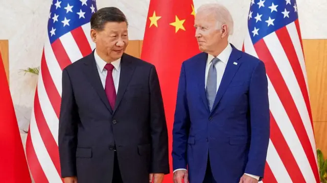 Xi Jinping e Joe Bidenbetway lplpébetway lplfrente a bandeiras, com sorrisos contidos