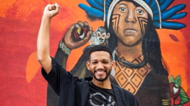 Rick Azevedo em frente a grafite, erguendo o braço e sorrindo 