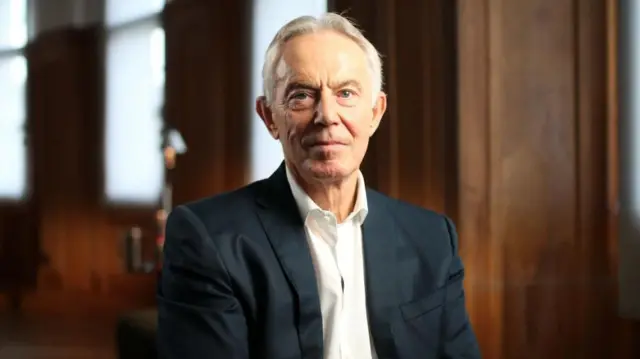 An image of former UK prime minister Tony Blair looking at the camera while dressed in a black suit and white shirt in a room with wooden walls. 