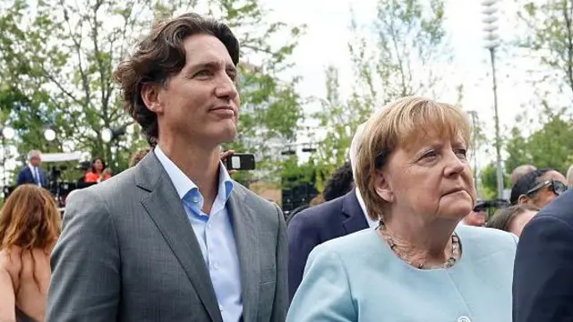 Justin Trudeau y Angela Merkel en la inauguración. Él viste traje gris con camisa clara y ella una chaqueta azul celeste